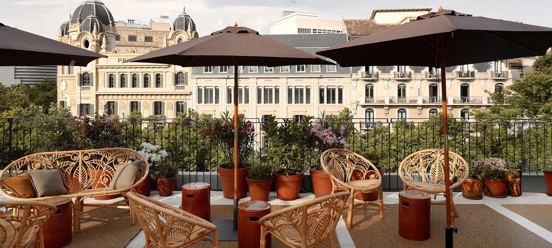 Rooftop terrace at Yurbban Ramblas with peacock-back rattan chairs and brown umbrellas, facing the domed towers of the Reial Academia de Ciencies building