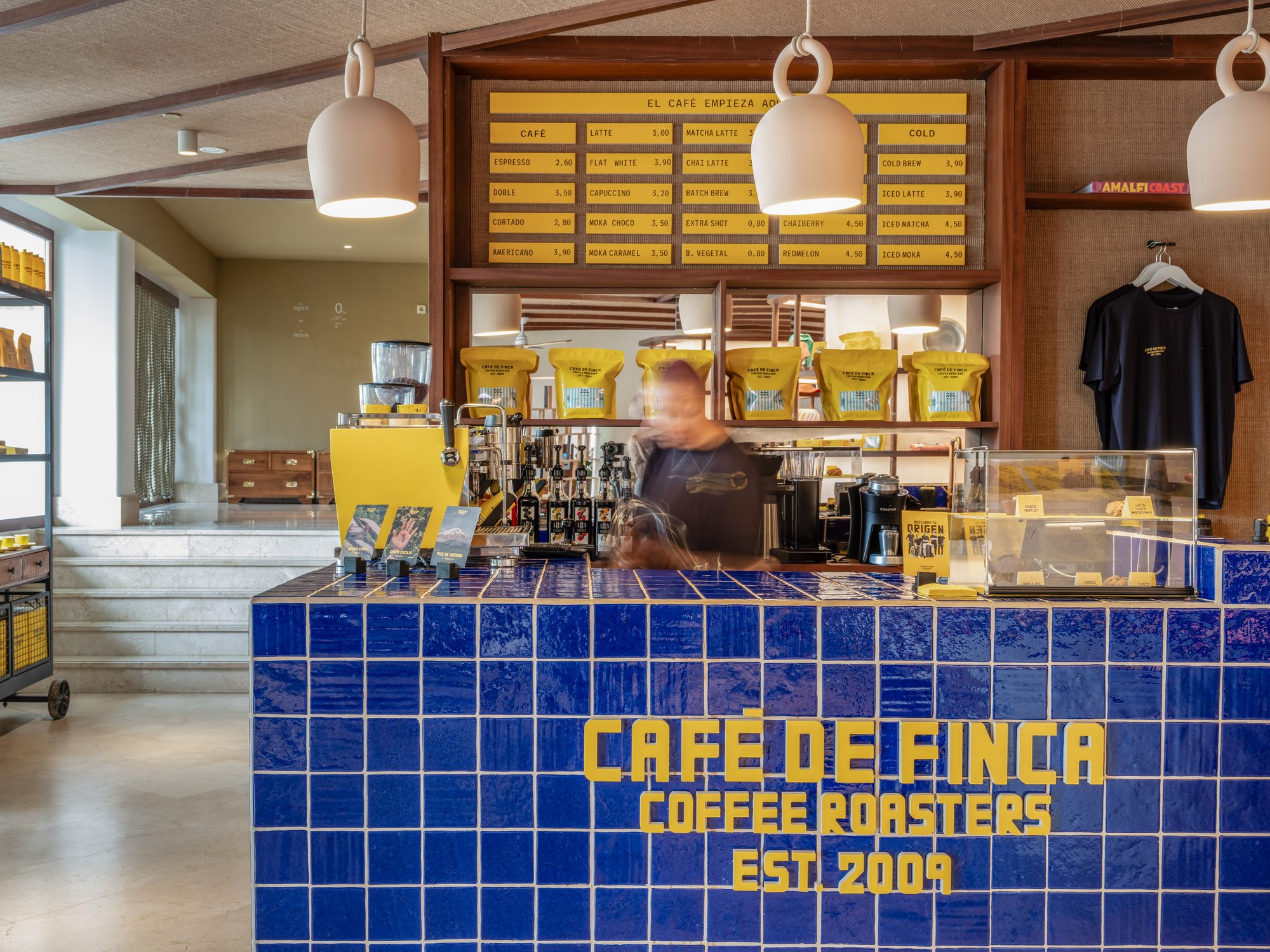 Cafe de Finca coffee bar at Zel Mallorca with a cobalt-blue tiled counter lettered "Cafe de Finca Coffee Roasters Est. 2009" and a barista working the espresso machine