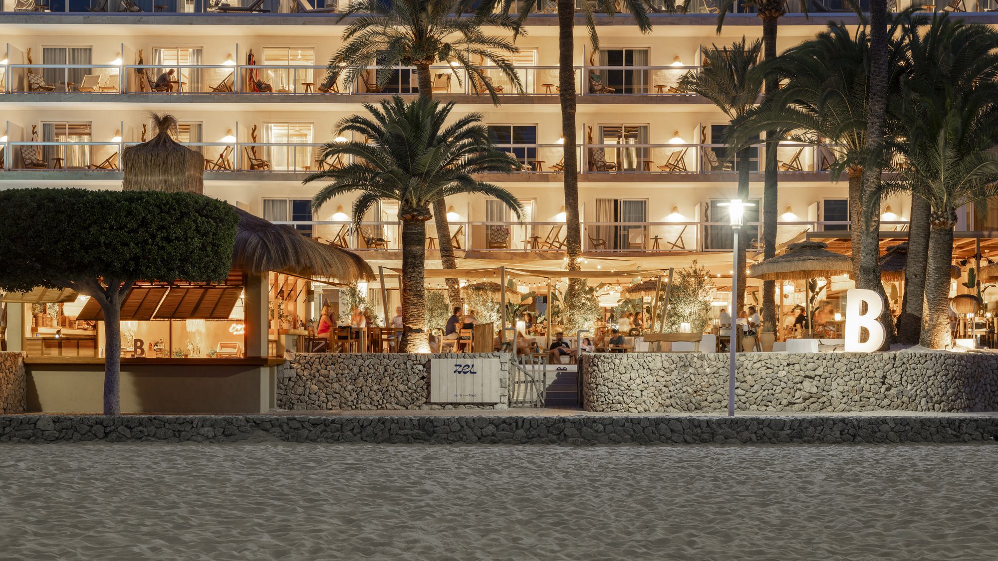 Night view of Zel Mallorca's beachfront facade from the sand, with lit balconies, palm trees, and a thatched beach bar marked with a large letter B
