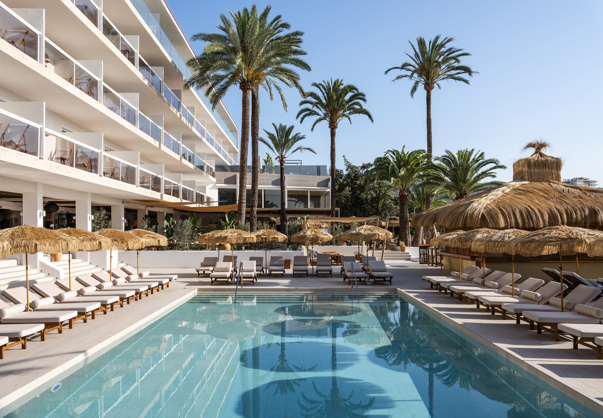 Main pool at Zel Mallorca lined with cream sun loungers and a thatched-roof cabana, facing the hotel's white balconied facade and palm trees