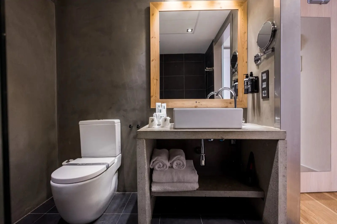 A hotel bathroom with a wood-framed mirror, a rectangular vessel sink on a concrete counter, rolled towels and Zenit-branded toiletries.