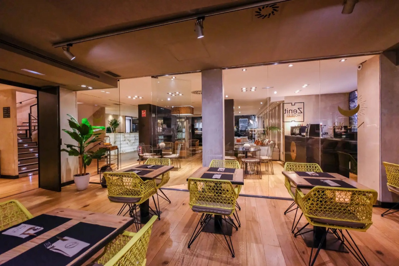 The hotel's restaurant and bar area with yellow woven chairs at set tables, a potted plant, and Zenit Hoteles signage on the back wall.