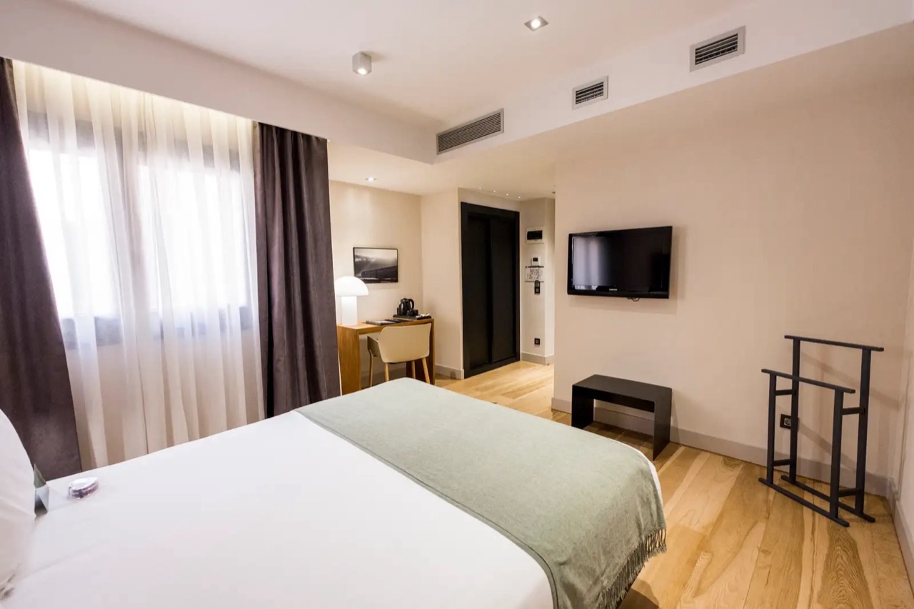 A hotel room with a bed dressed in white linen and a green throw, wood flooring, and a desk area beyond it.