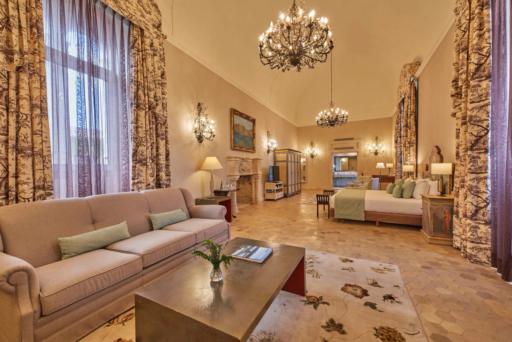 High-ceilinged elegant suite with a crystal chandelier, patterned drapery, a seating area, and a four-poster-style bed
