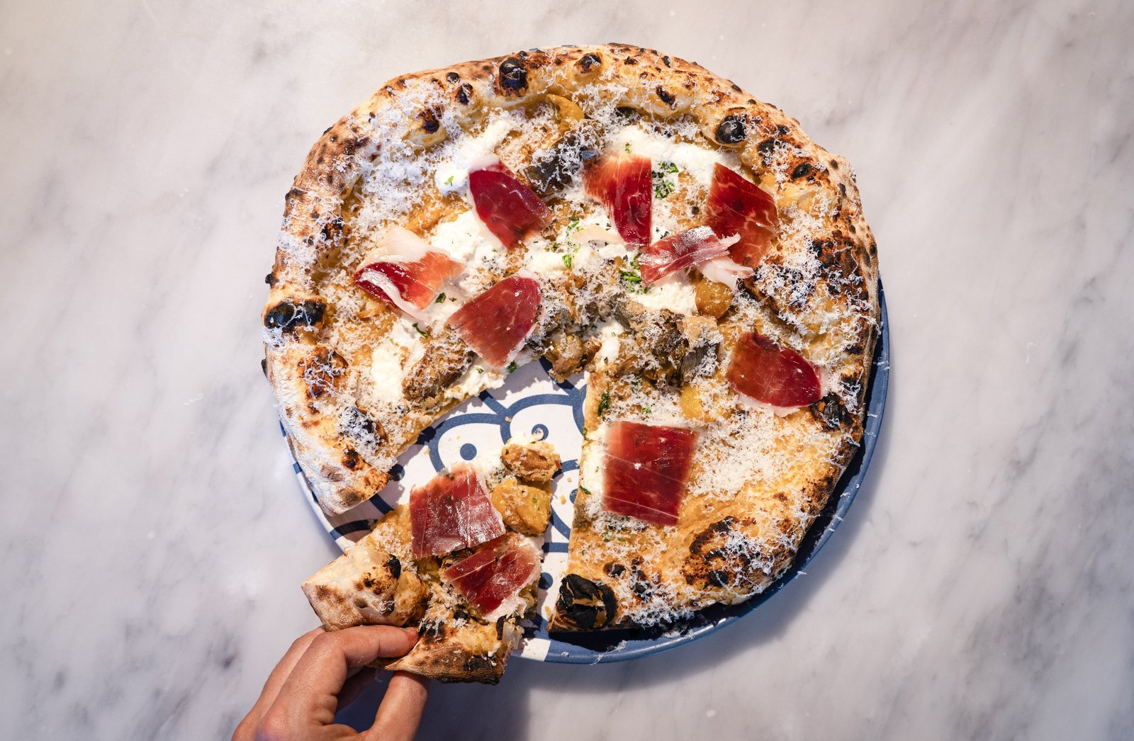 A Neapolitan-style pizza topped with cured ham on a marble table, with a hand lifting a slice.