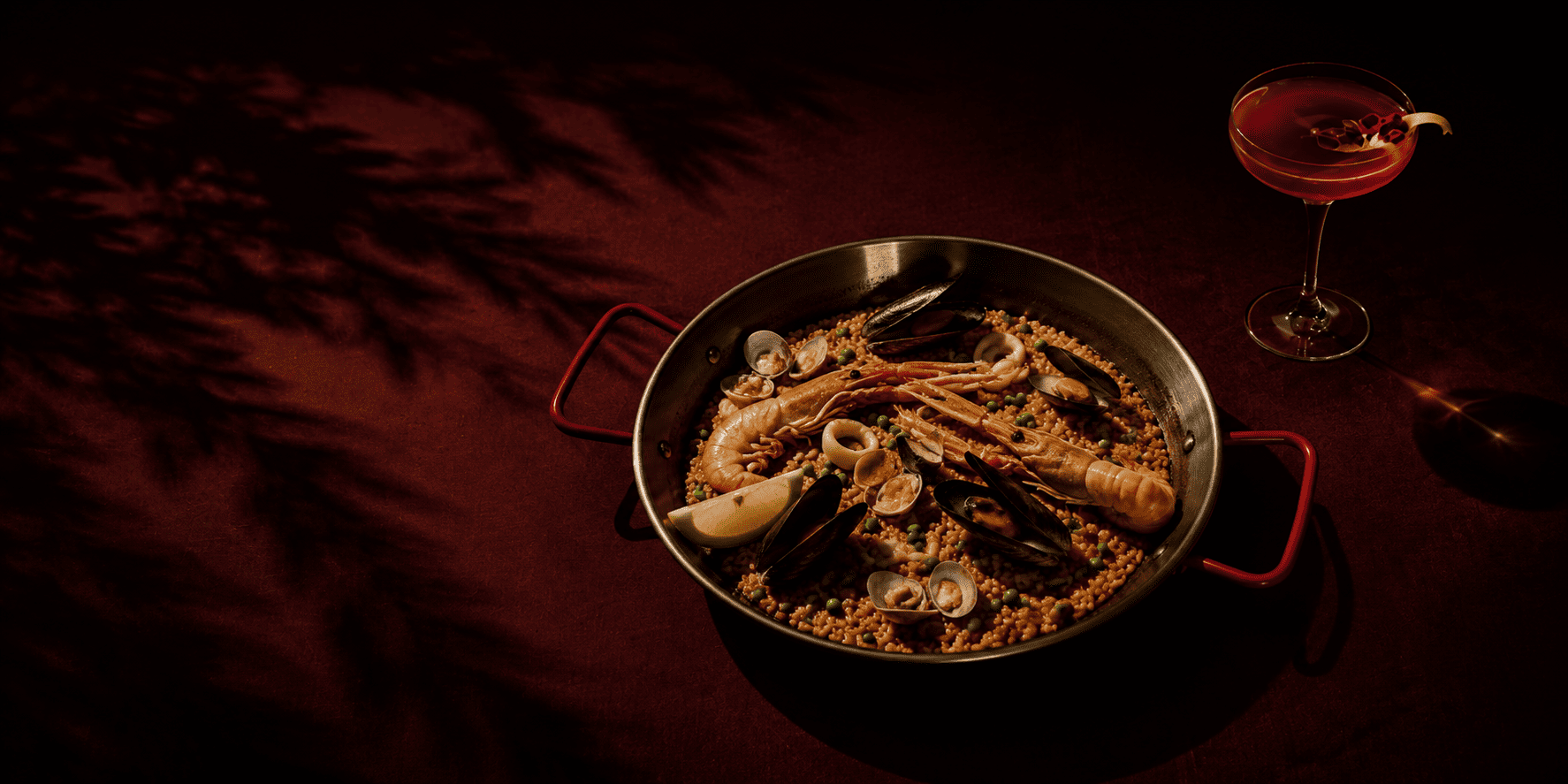 A seafood paella pan with prawns, mussels and clams beside a red cocktail on a dark table, moody low lighting, at Dalirium in Barcelona.