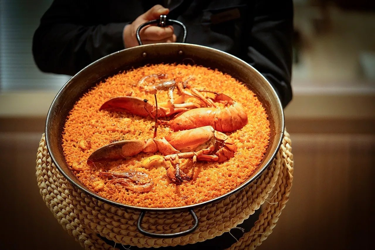 A waiter holding a paella pan of lobster rice resting on a woven stand at L'Albufera