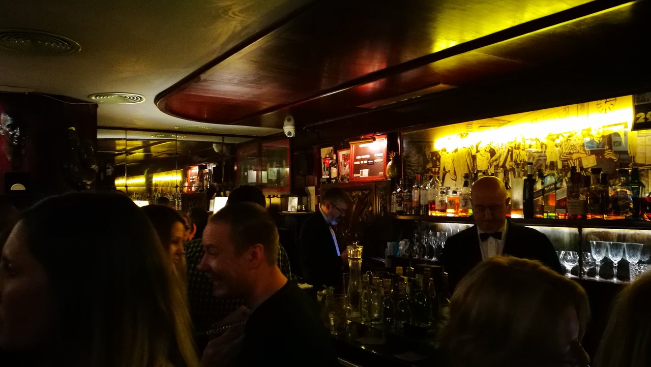 A bartender in a white shirt, black waistcoat and bow tie works behind the counter at Boadas Cocktail Bar in Barcelona, with backlit shelves of spirits and customers gathered at the bar in the foreground.