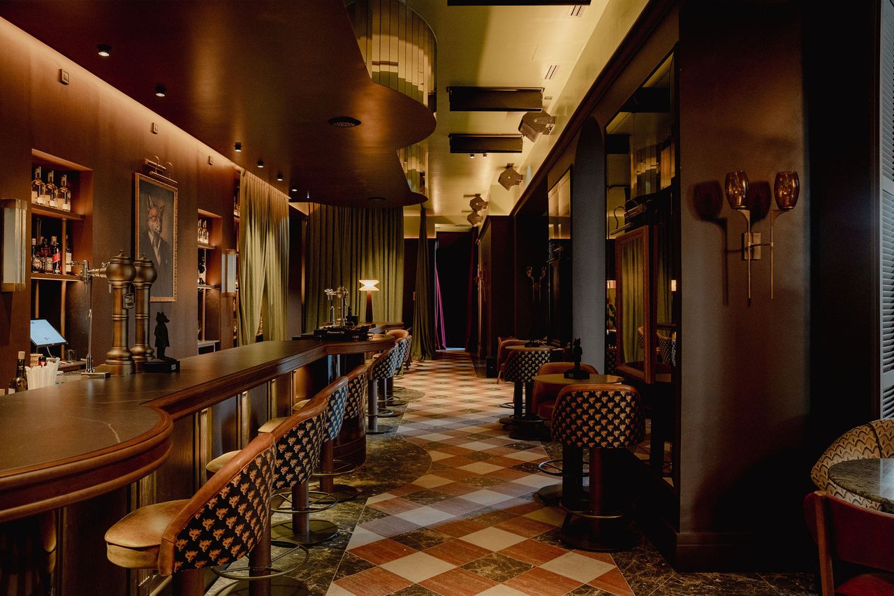 A long dark-wood bar with patterned stools, a fox portrait on the wall and a checkered floor at La Casa de Fox