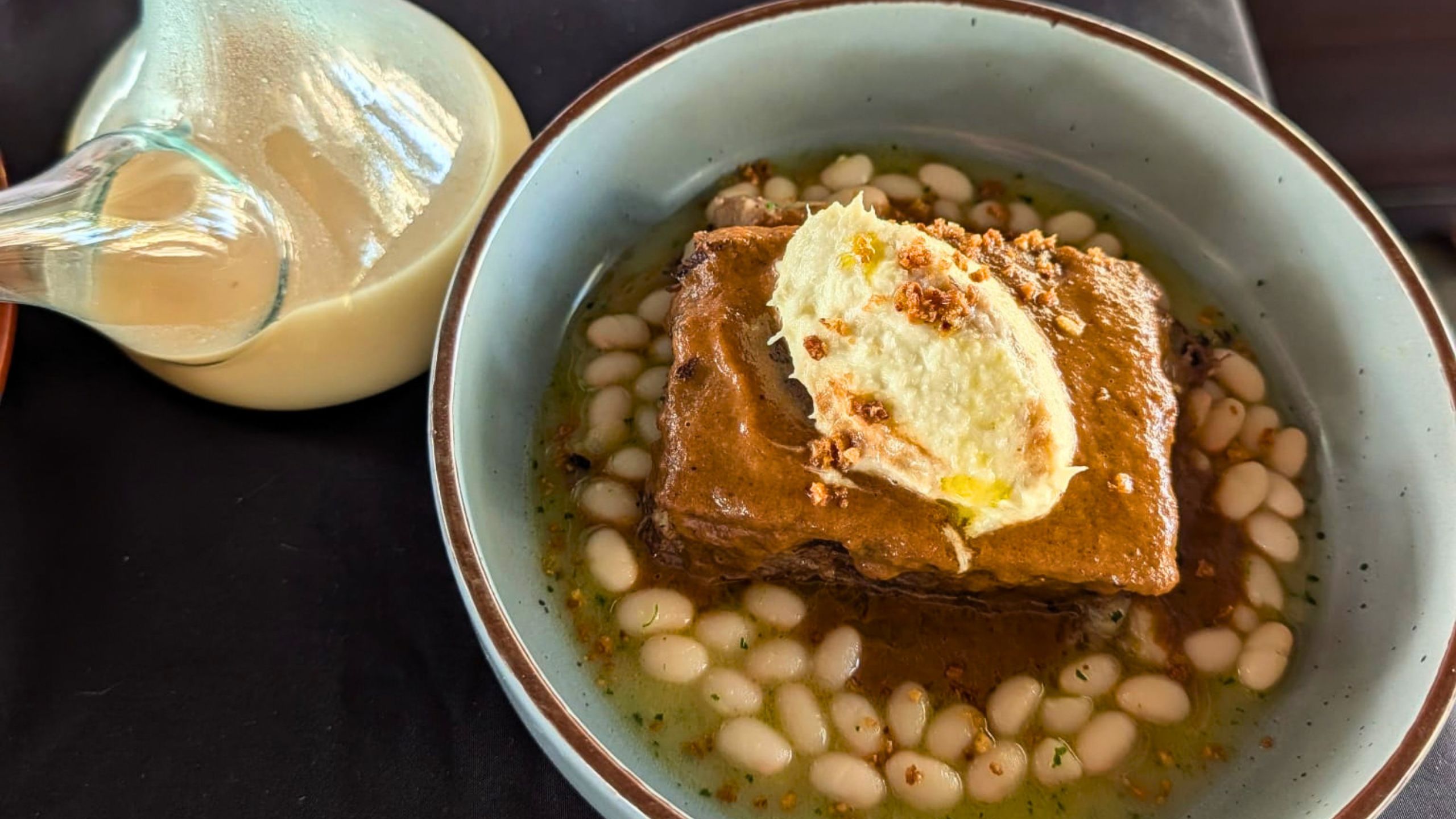 A bowl of white beans in broth topped with braised pork cheek and salt cod brandada, with a glass porró of bean broth alongside, L'Andreuenc's winning esmorzar de forquilla dish