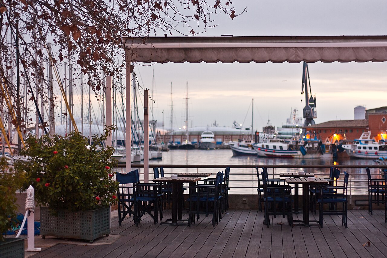 An empty restaurant terrace with tables and chairs under an awning overlooking the harbour at the Port of Palma, Mallorca.