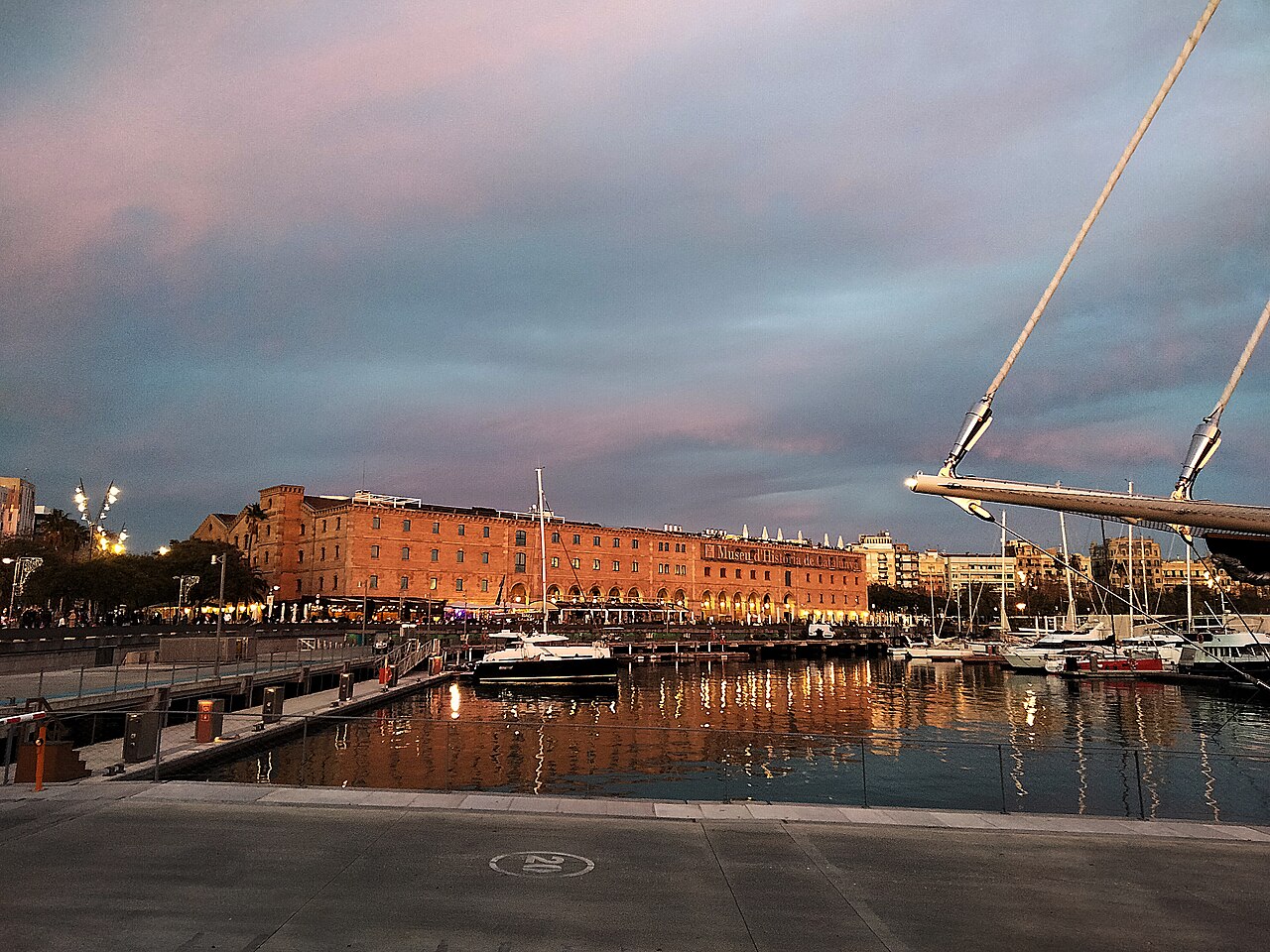 Dusk over the Port Vell marina in Barcelona, with the brick Palau de Mar building lit up across the water (illustrative, the restaurant itself is not shown)