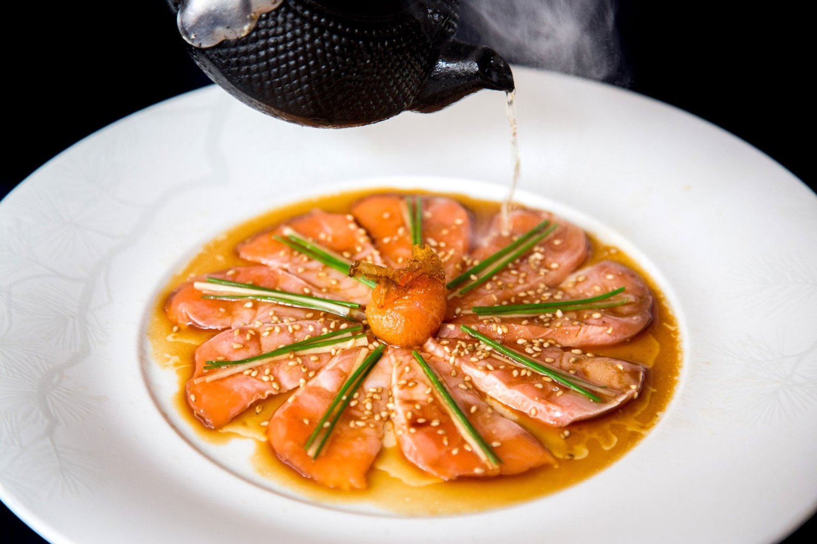 Slices of fish arranged in a circle in a white bowl as sauce is poured over them.