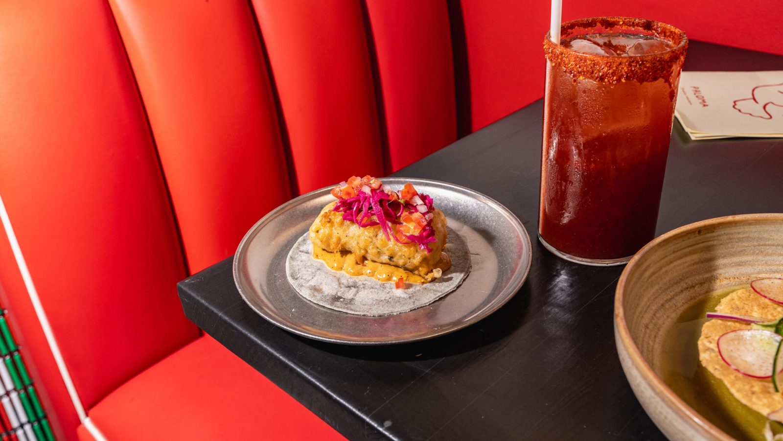 A fish taco topped with pickled red onion beside a chilli-rimmed drink on the red booths at Paloma Fondita Mexicana.
