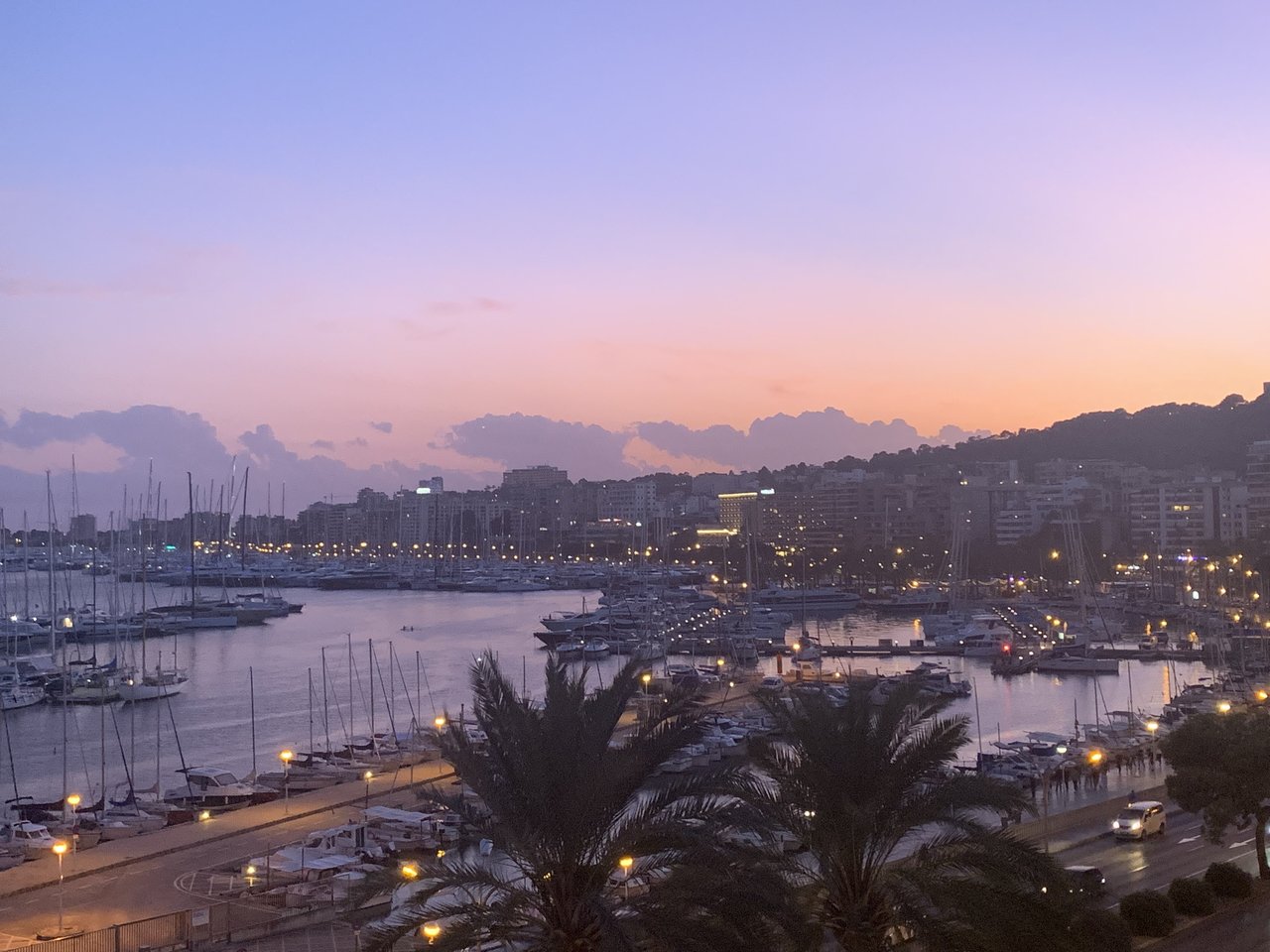 Sunset over Palma de Mallorca's marina, with moored boats, palm trees and the city skyline along the Passeig Marítim.