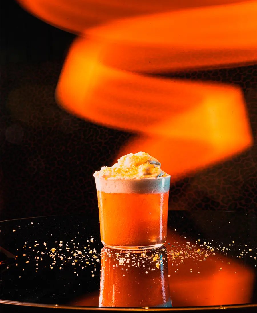 An orange cocktail on a dark bar, lit from behind by a curl of orange light.