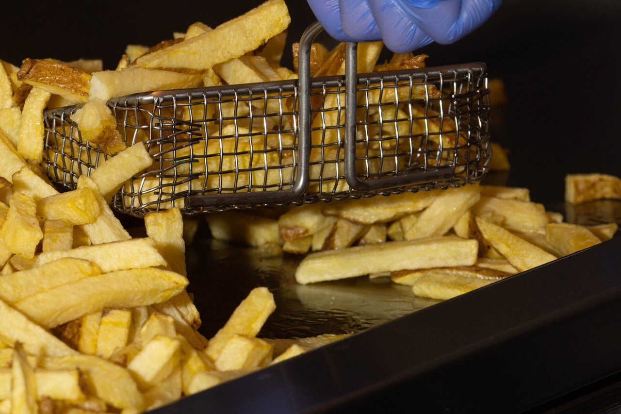 A gloved hand lifting a wire basket of freshly fried chips from a tray of fries