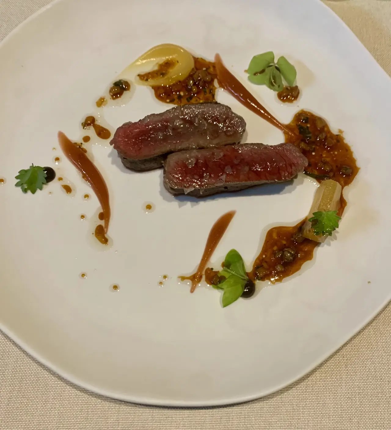 Two slices of seared, pink-centred meat with dots of sauce and a green garnish on a white plate.