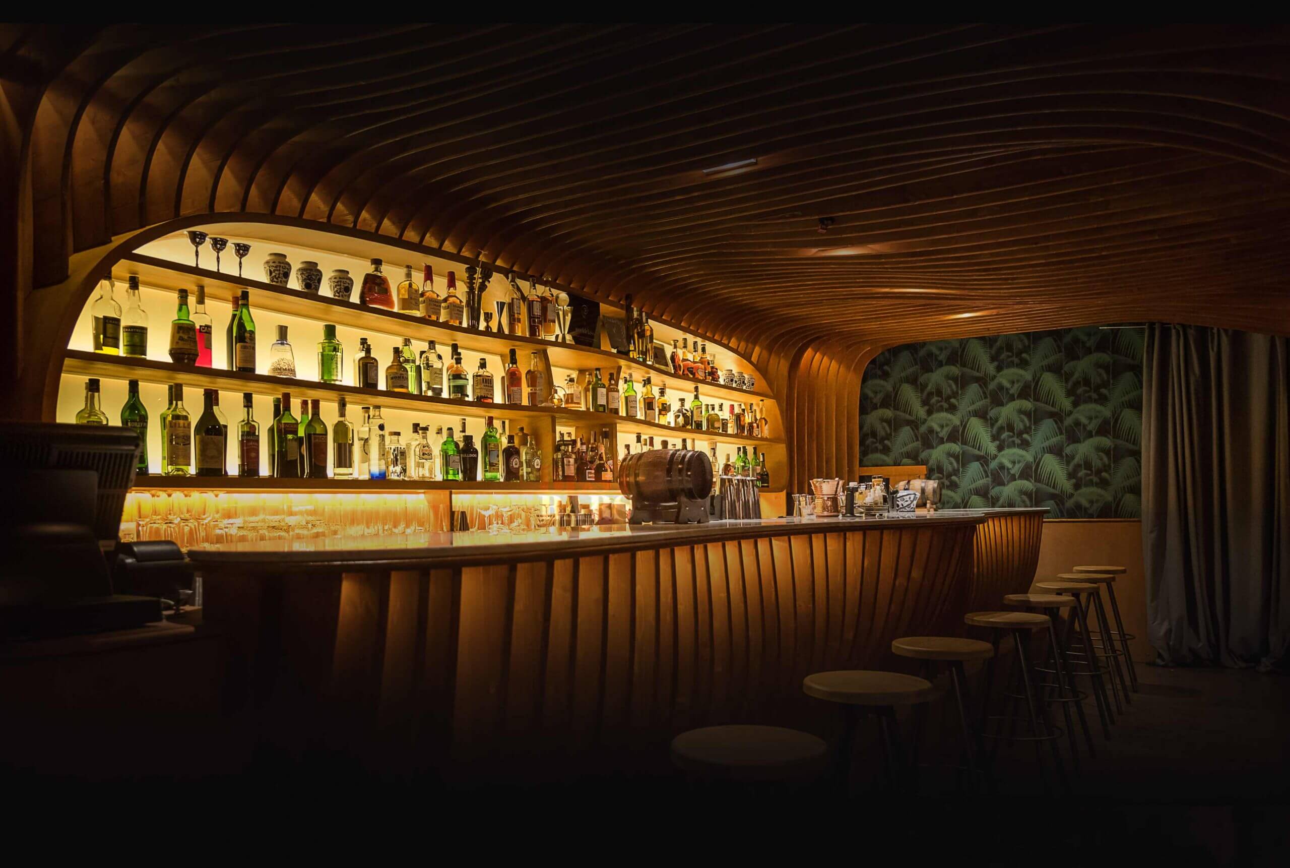 The curved wooden bar and ribbed timber ceiling at Paradiso in Barcelona, with bottles lit on shelves behind the counter and stools along the bar