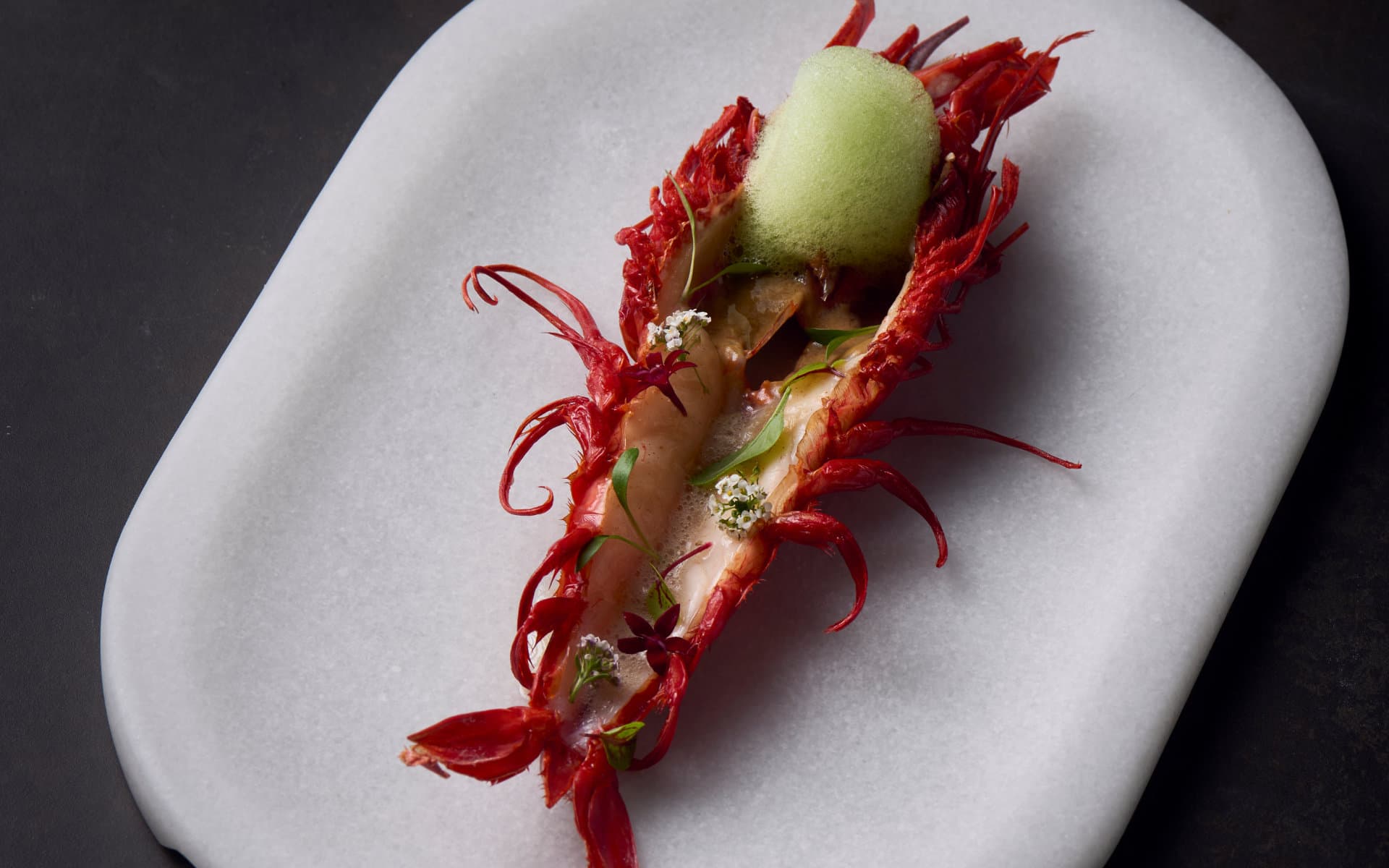 A whole red prawn plated on a white marble dish with a scoop of green apple sorbet, edible flowers and herb leaves, at Slow & Low in Barcelona.