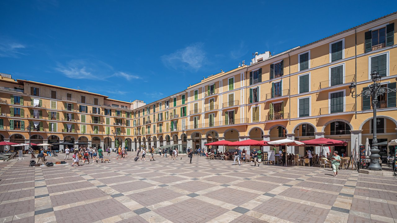 Restaurant terraces with red and white umbrellas on Plaça Major in Palma de Mallorca, with people walking across the plaza.