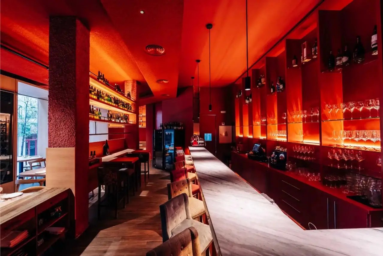 The long bar at Angelita, lit red, with stools along the counter and bottles on shelves behind