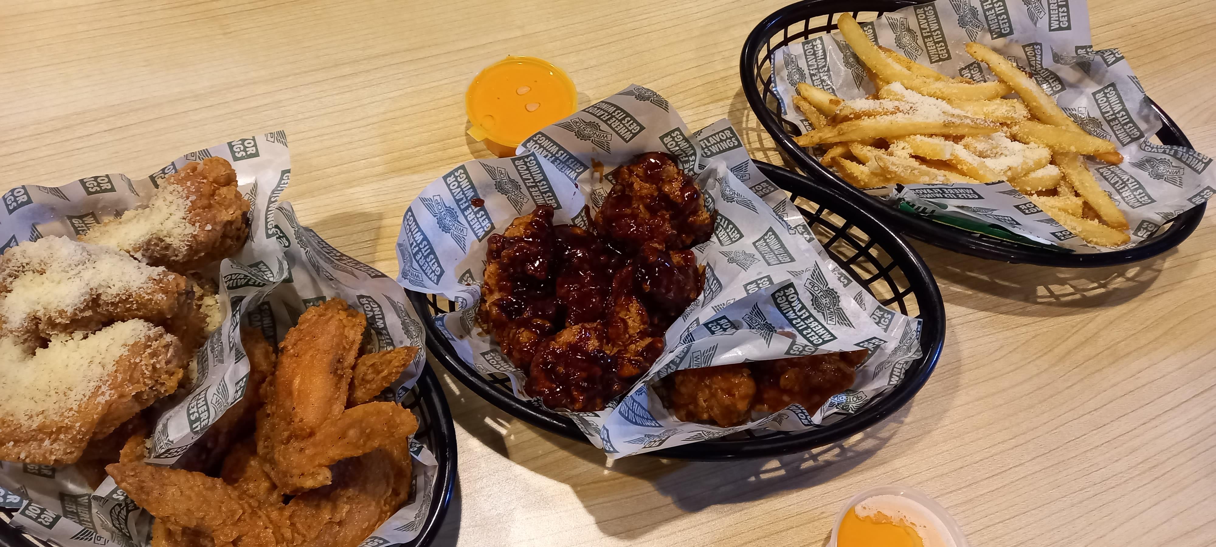 Baskets of Wingstop fried chicken tenders, saucy boneless wings and parmesan-garlic seasoned fries on Wingstop-branded wax paper, with small cups of dipping sauce on a wood-look table.