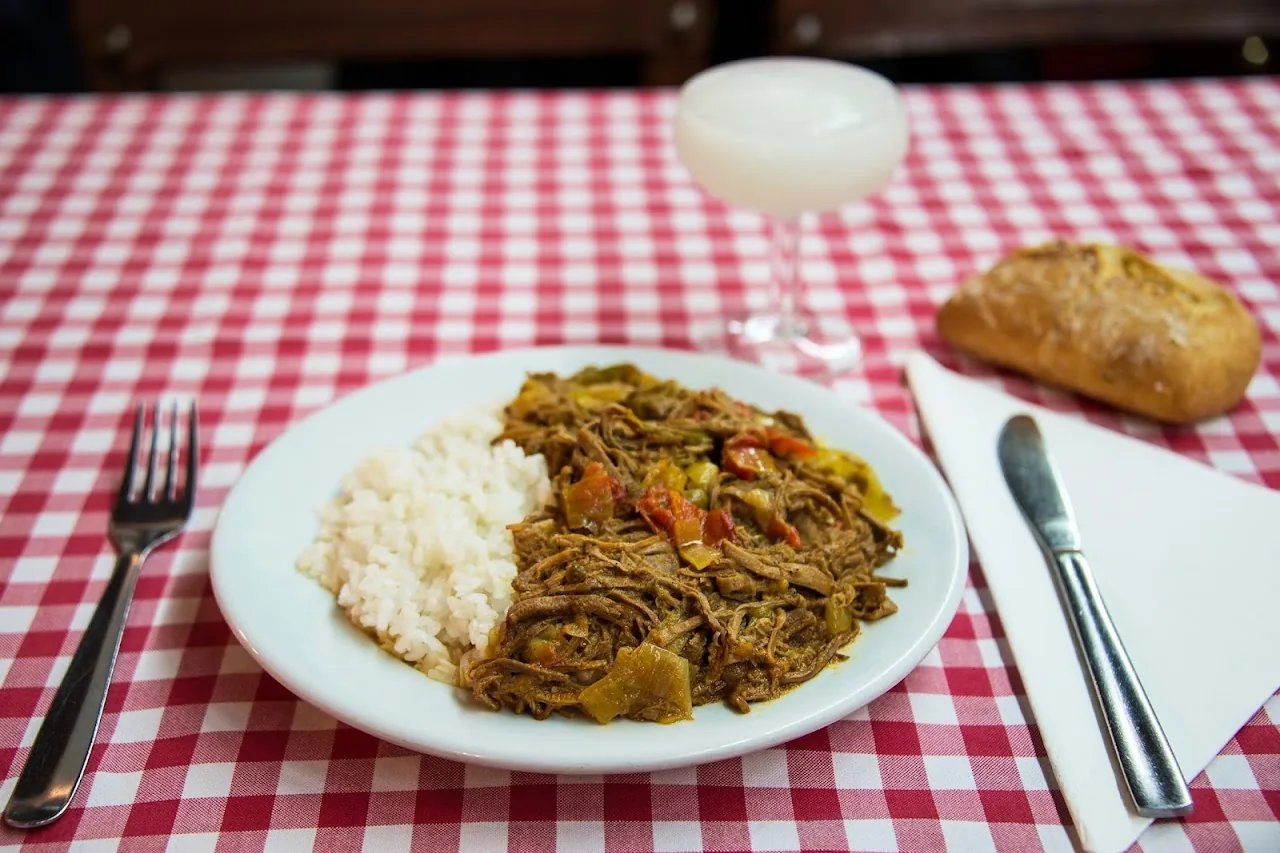 A plate of shredded ropa vieja beef with white rice on a red-checked tablecloth, with bread and a cocktail behind
