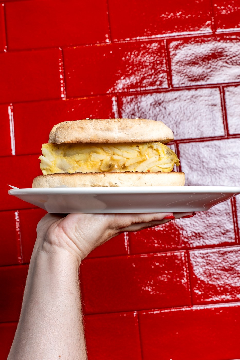 A hand holding a plate with a grilled llonguet filled with thick Spanish potato omelette, against a red-tiled wall.